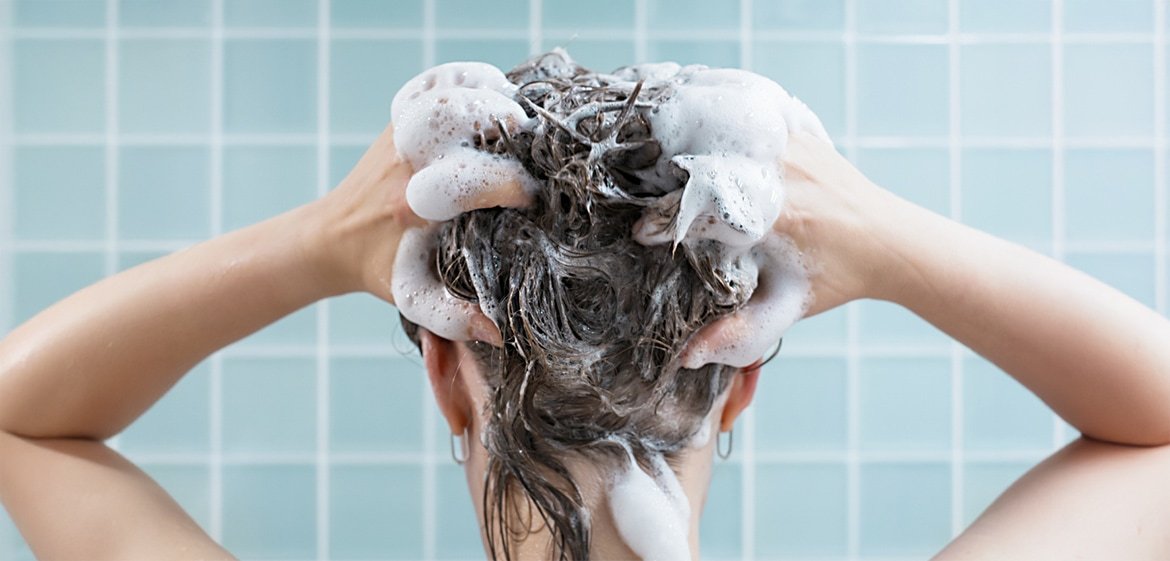 Der Kopf einer Brünetten von hinten. Sie shampooniert sich mit beiden Händen die Haare ein.